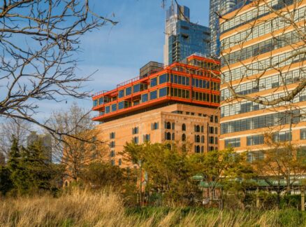 A multi-story brick building with a modern, biomimicry-inspired orange rooftop addition, viewed behind trees and tall grass.