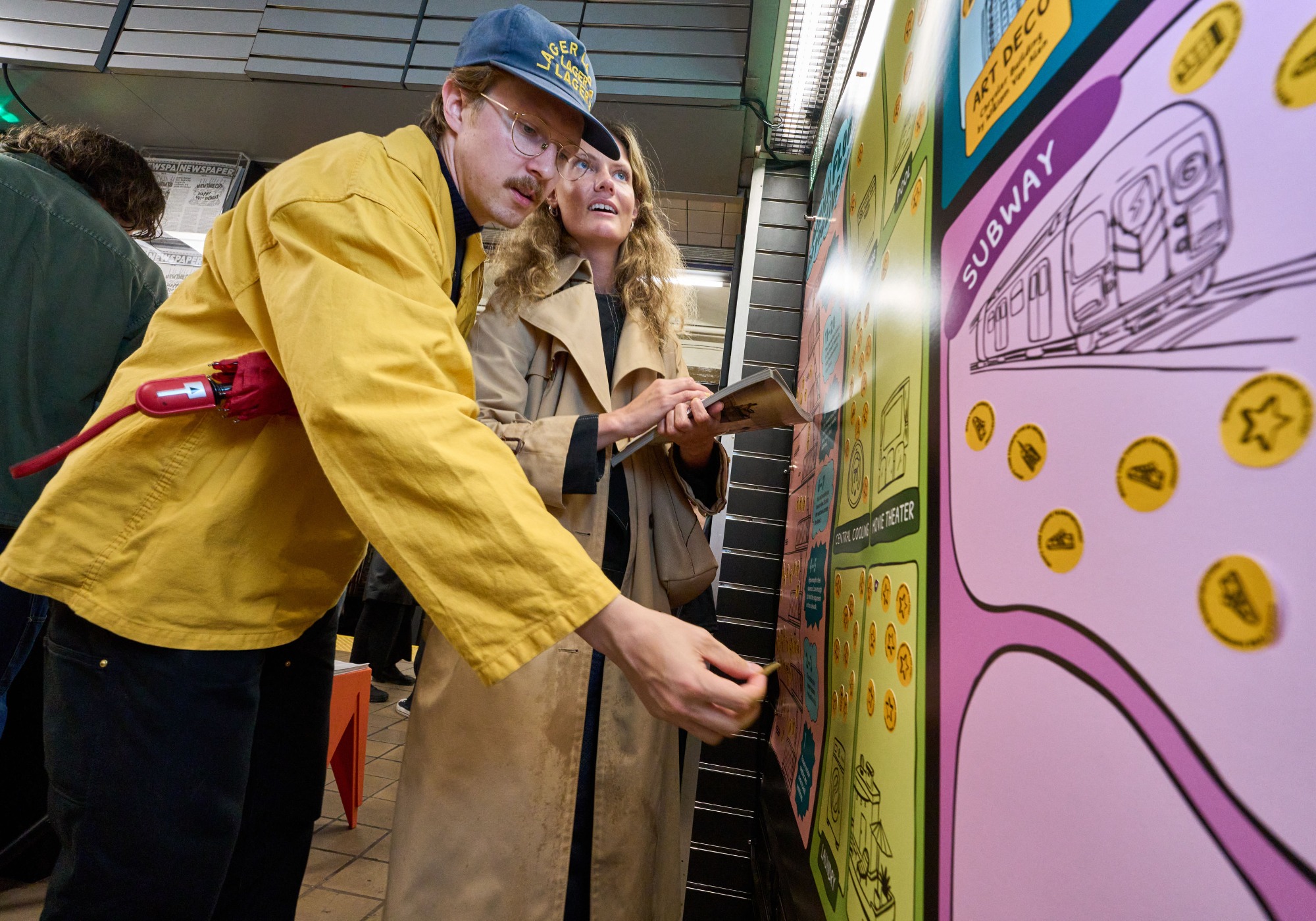 Two people in coats interact with a colorful wall display featuring subway art and tokens inside a subway station during Archtober 2026.
