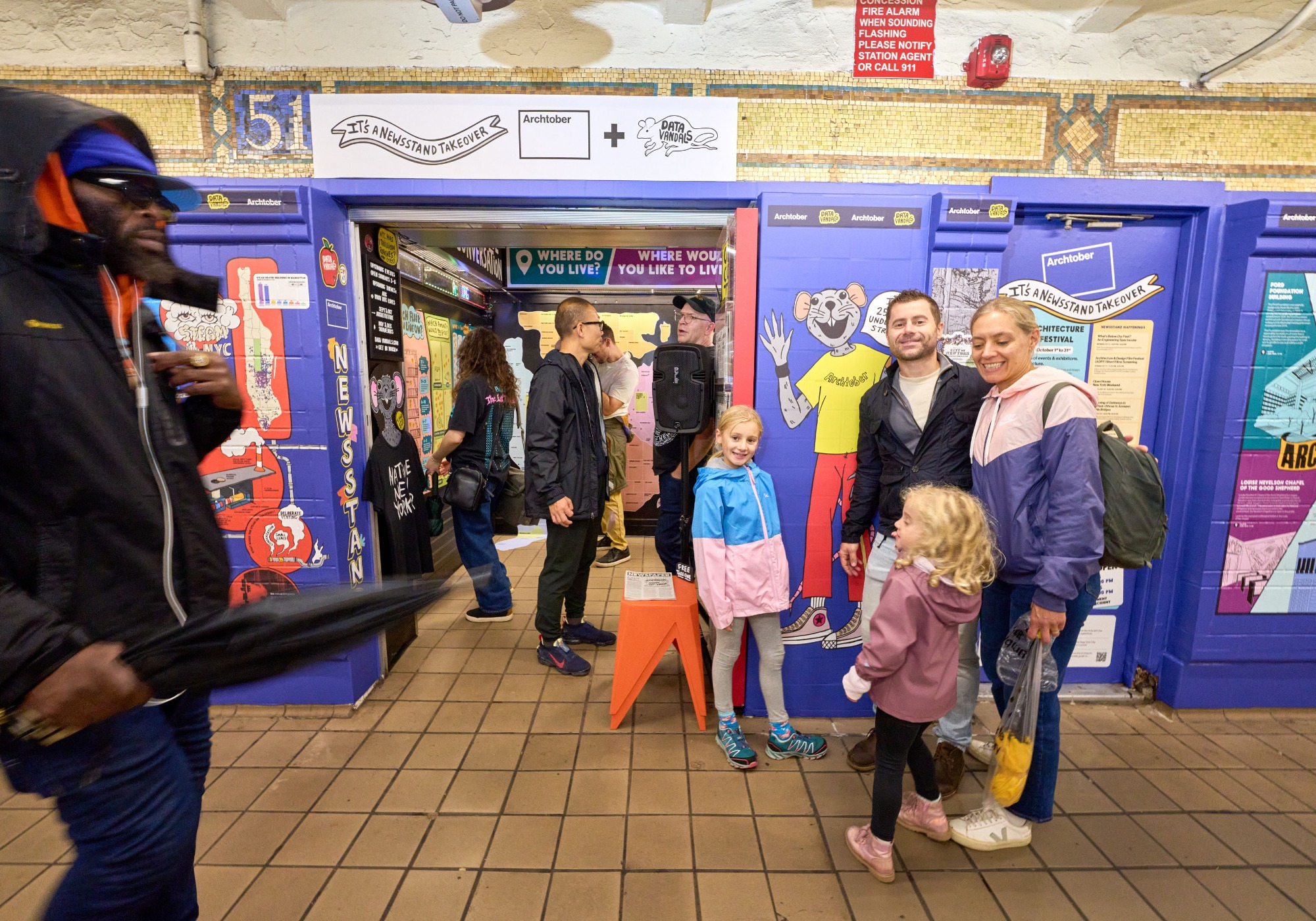 People stand and walk inside a subway station with colorful murals; during Archtober 2026, a family poses as others pass by. The scene appears lively and well-lit.