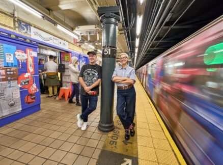 Two people stand on a subway platform near a “51” pillar during Archtober 2026, with a blurry train passing by and others using a ticket machine in the background.