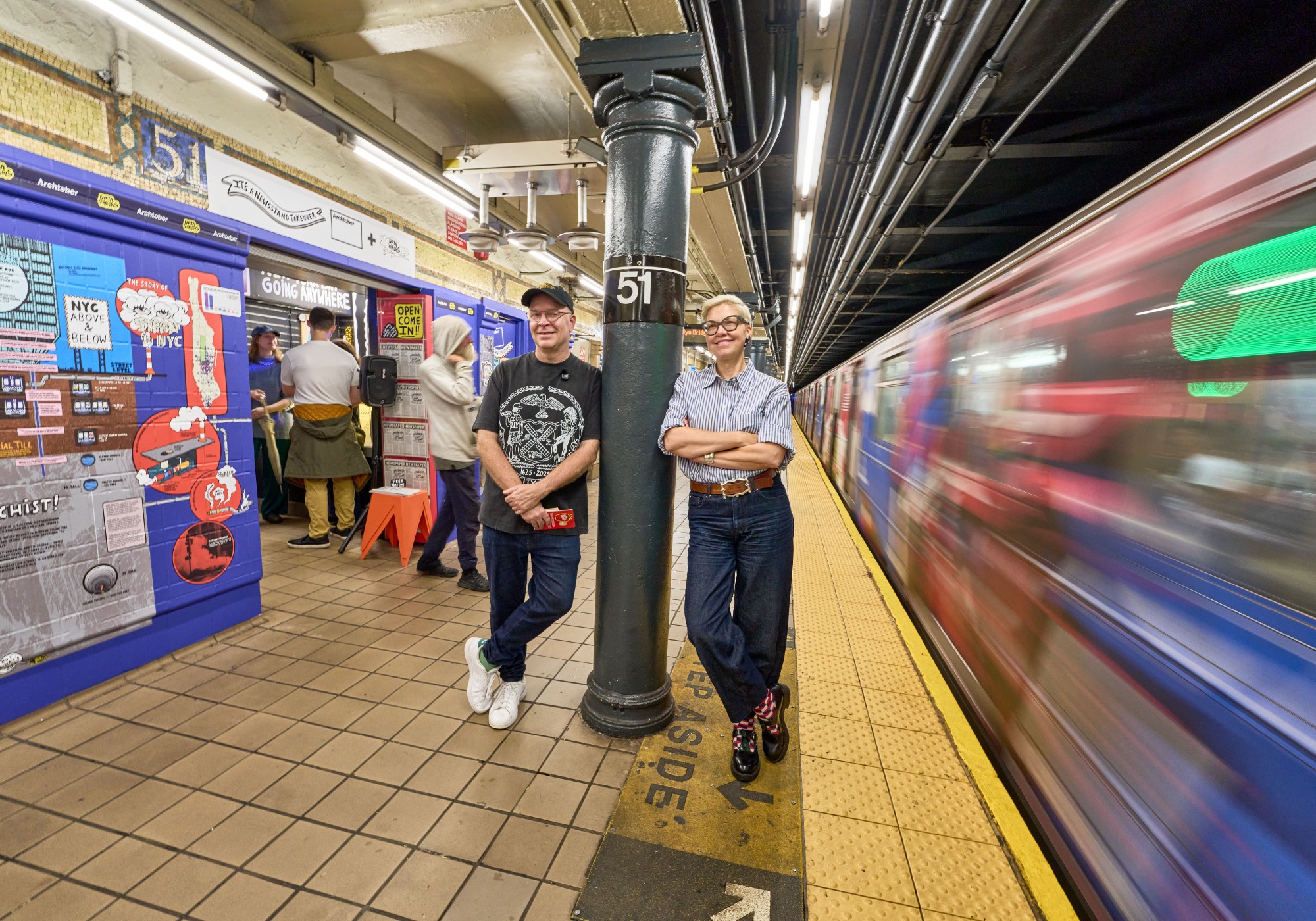 Two people stand on a subway platform near a “51” pillar during Archtober 2026, with a blurry train passing by and others using a ticket machine in the background.