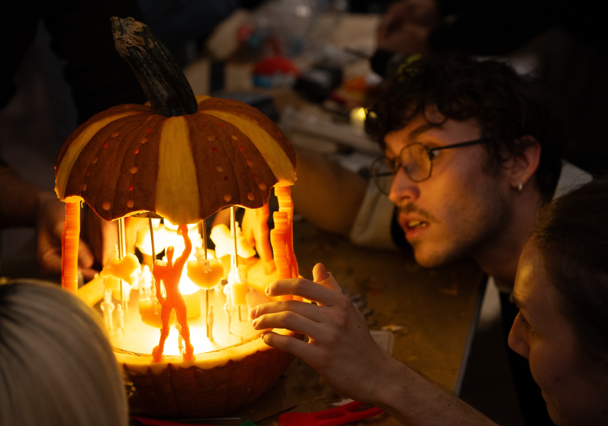 A person examines a carved pumpkin lit from within, featuring detailed miniature figures and columns, resembling a glowing carousel.