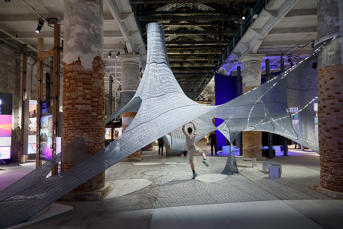 A person jumps under a large, white, mesh-like architectural installation at MIT Future Fest inside an industrial brick hall.