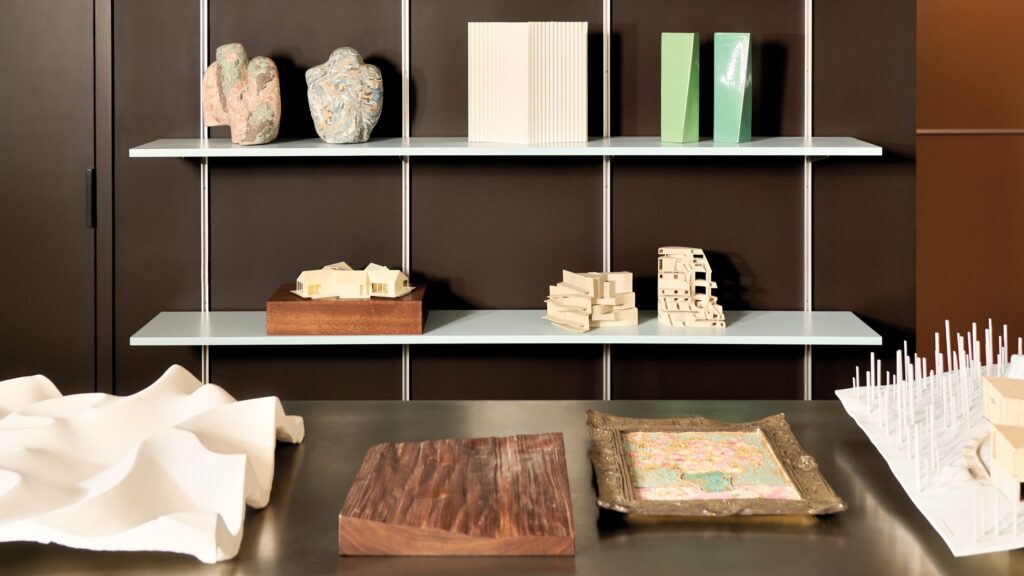 Two shelves holding various abstract sculptures and architectural models, with additional models, a wood block, and a framed painting on the table below.