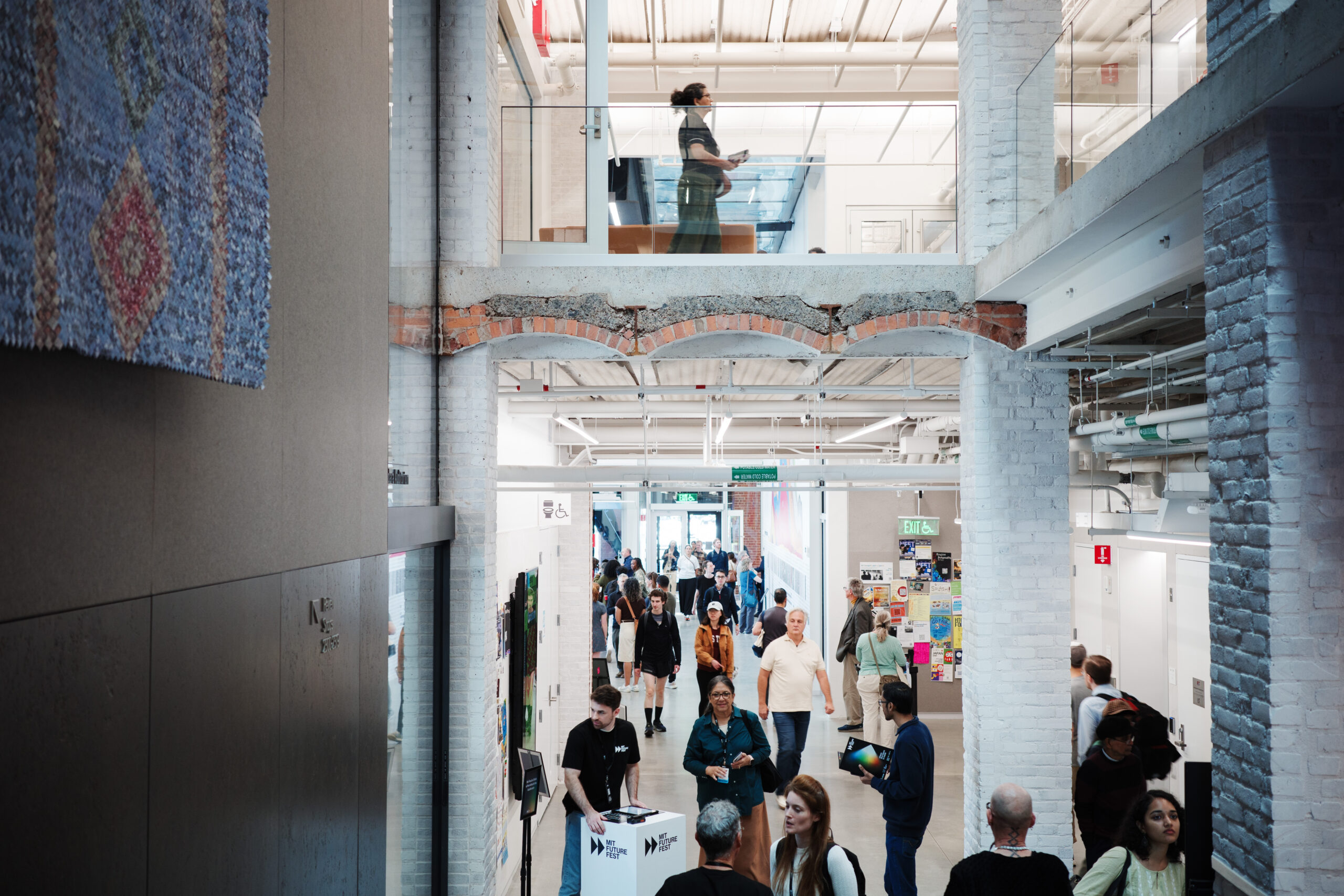 People walk through the central hallway of an MIT Future Fest multi-level building as another person stands on an upper-level mezzanine.