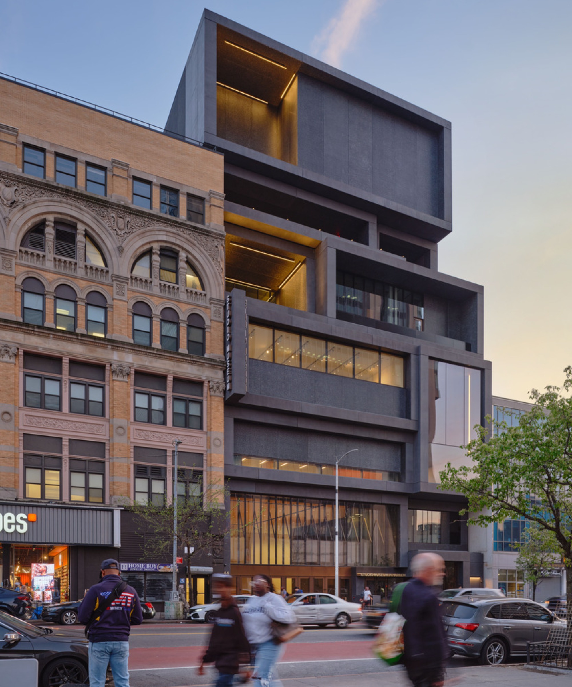 A modern, multi-story black building with geometric design stands beside an ornate brick structure as people stroll by, with cars parked along the street.