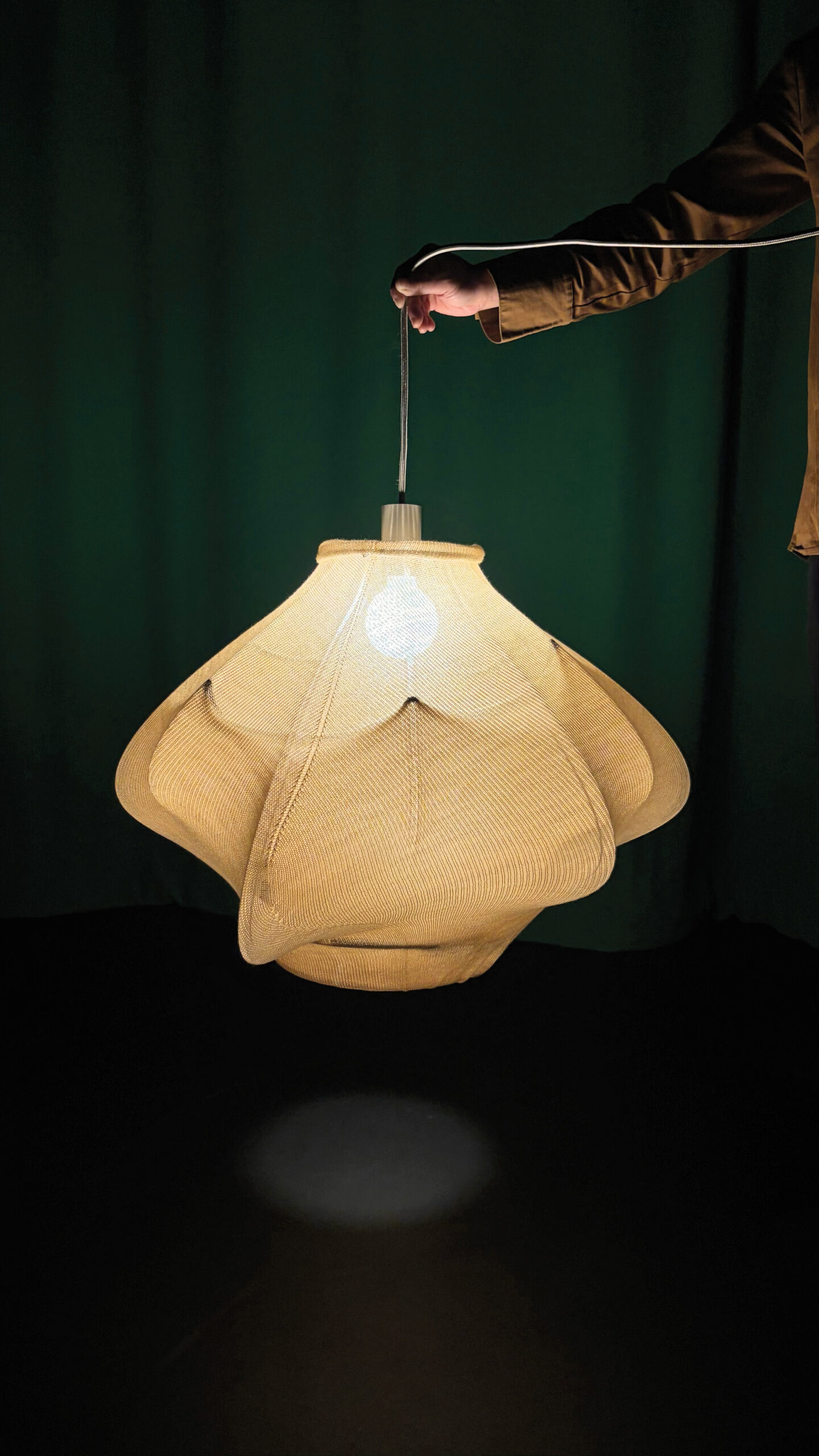 Made by Yu Nong Khew, a hand holds up a beige, angular pendant light with a lit bulb inside.