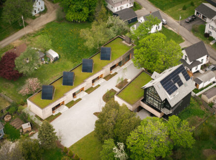 Aerial view of a residential property featuring two modern buildings with green roofs and solar panels, surrounded by a gravel driveway and lawn.