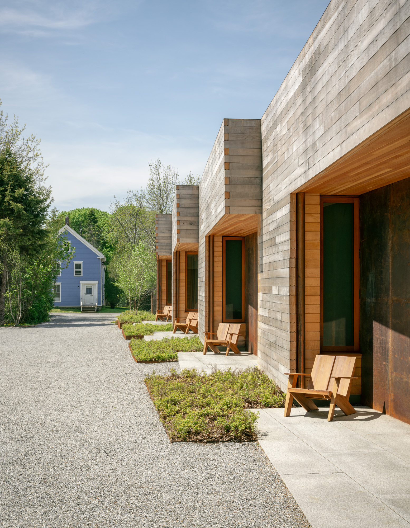 A row of modern wooden cabins at the Ellis-Beauregard Foundation with small patios and chairs, adjacent to a gravel path and a blue house.