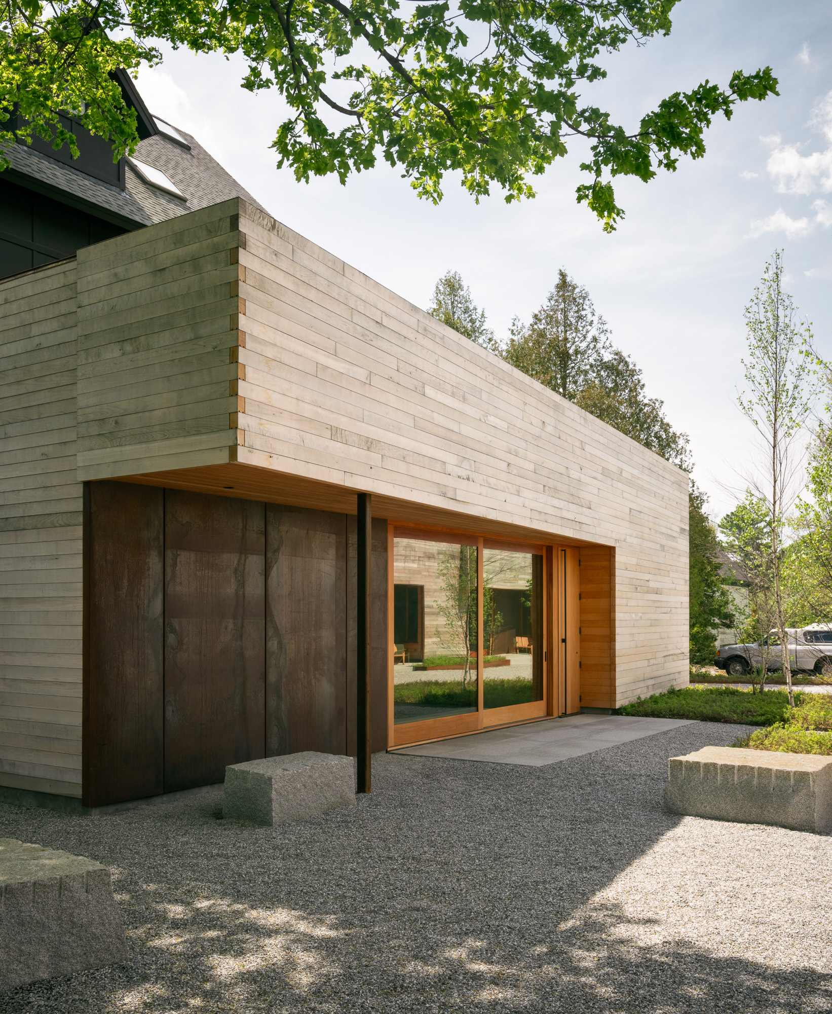 Modern Ellis-Beauregard Foundation house exterior featuring horizontal wood siding, a dark metal panel wall, and large glass sliding doors, set beside a gravel courtyard with stone blocks.