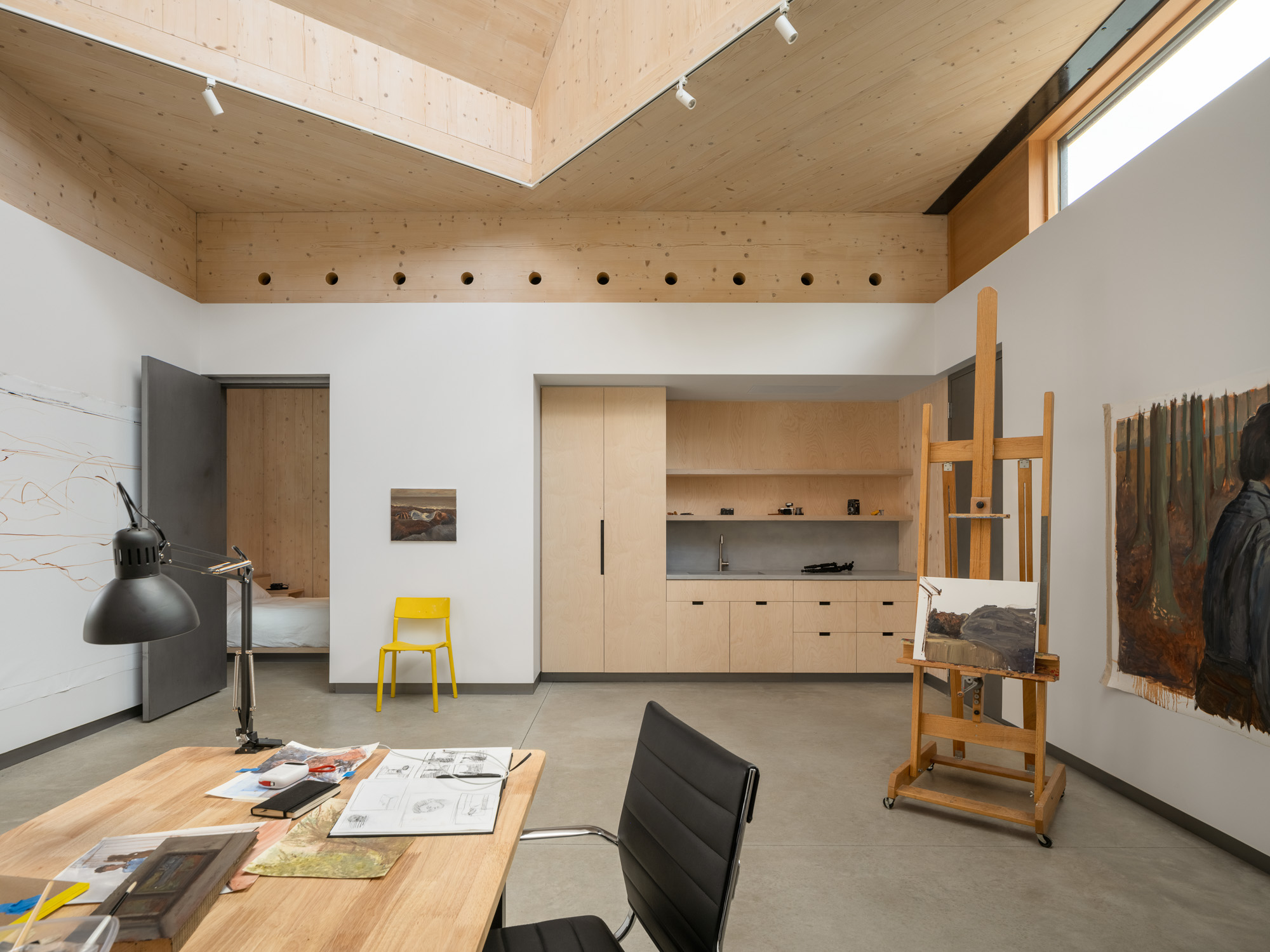 A studio space at the Ellis-Beauregard Foundation with a desk in the foreground, a yellow chair, a wooden easel holding a painting, and light wood cabinetry with a sink against the back wall.