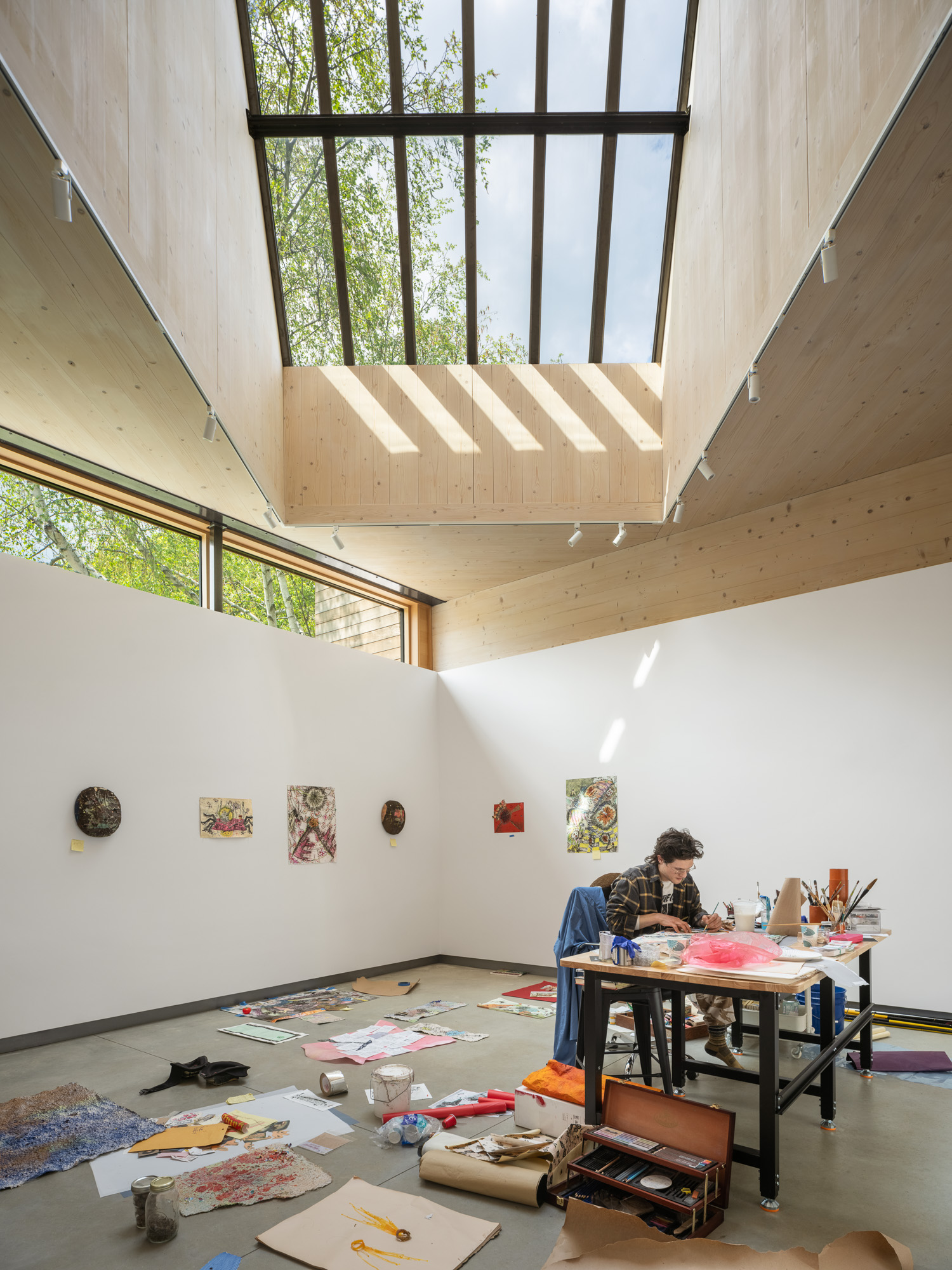 A person works at a desk in an Ellis-Beauregard Foundation studio with a high ceiling, a large skylight, and various artworks displayed on the walls.