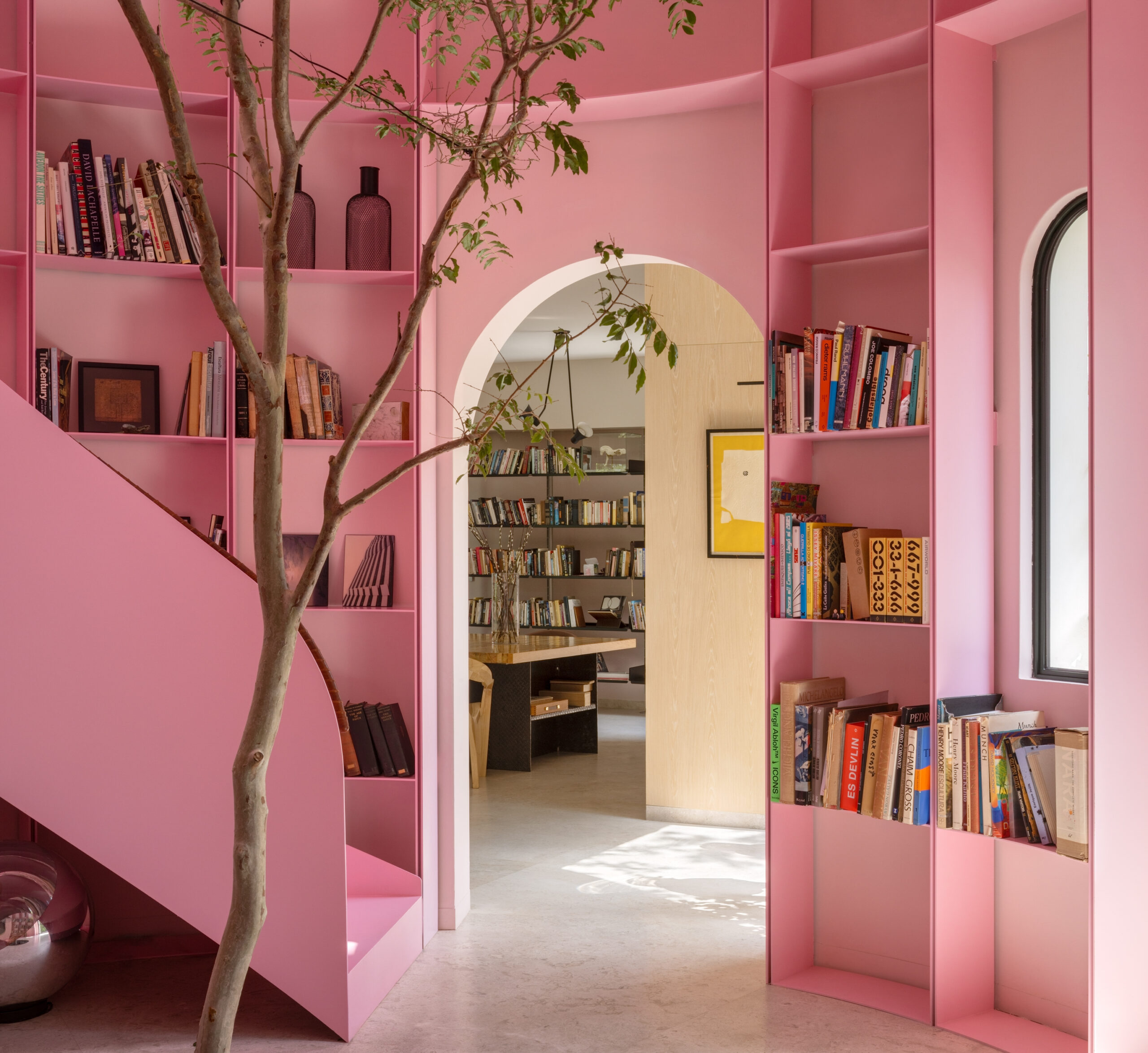 A room with floor-to-ceiling pink bookshelves, a central staircase, and a small tree, viewed through an arched doorway leading to another library space.