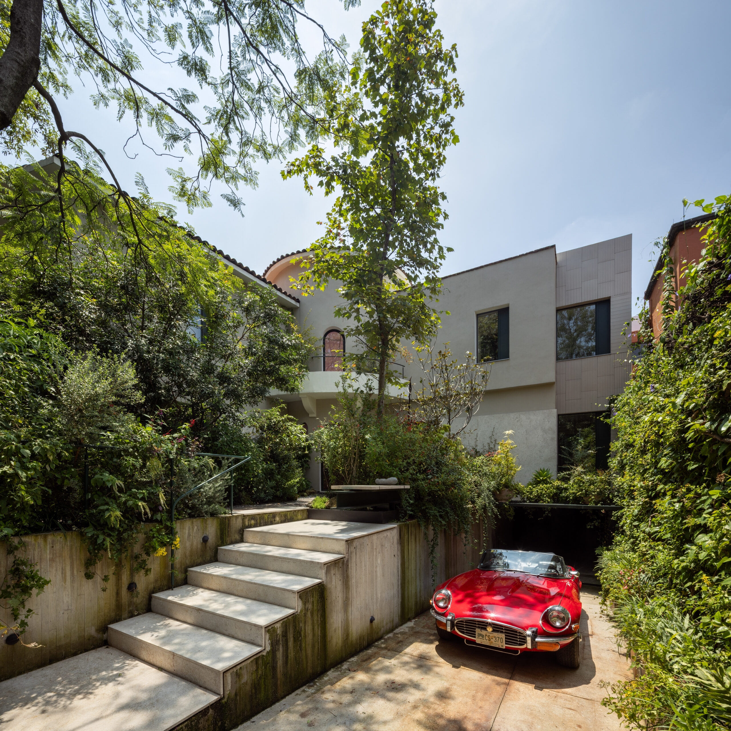 A red convertible sports car parked in the driveway of a modern home with concrete steps and lush landscaping.
