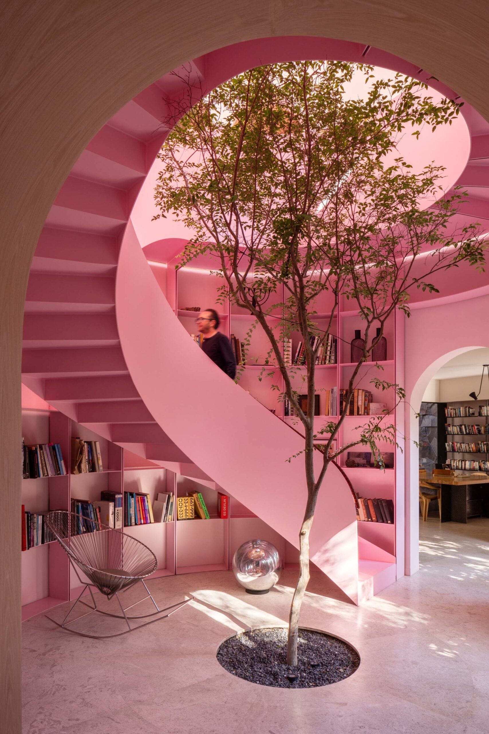 A person walking up a bright pink spiral staircase that winds around a small indoor tree.