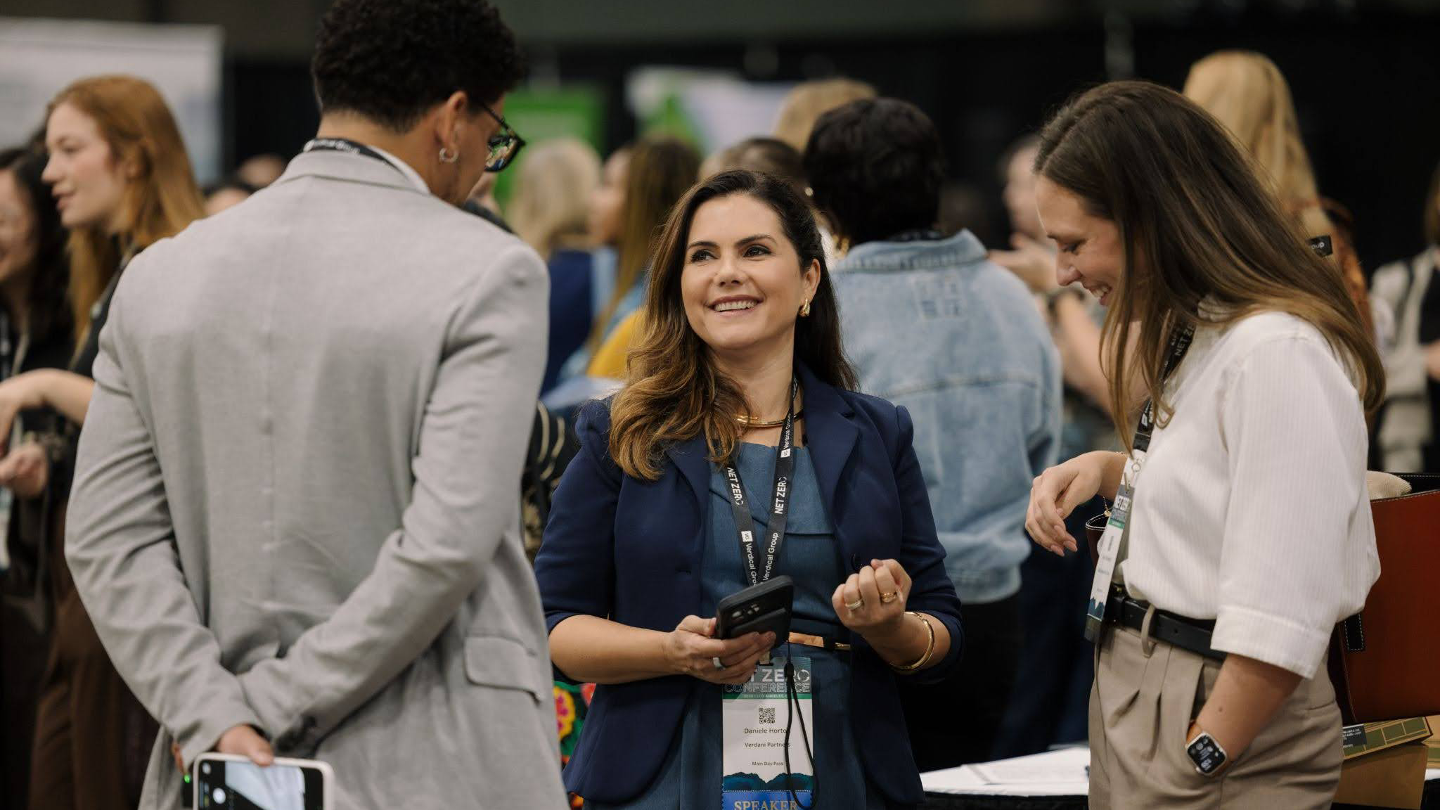 A woman wearing a navy blazer and lanyard smiles while talking to two people at a conference.