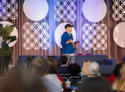 A man in a blue hoodie and baseball cap speaks on stage before an audience, with geometric wall decorations and plants in the background.