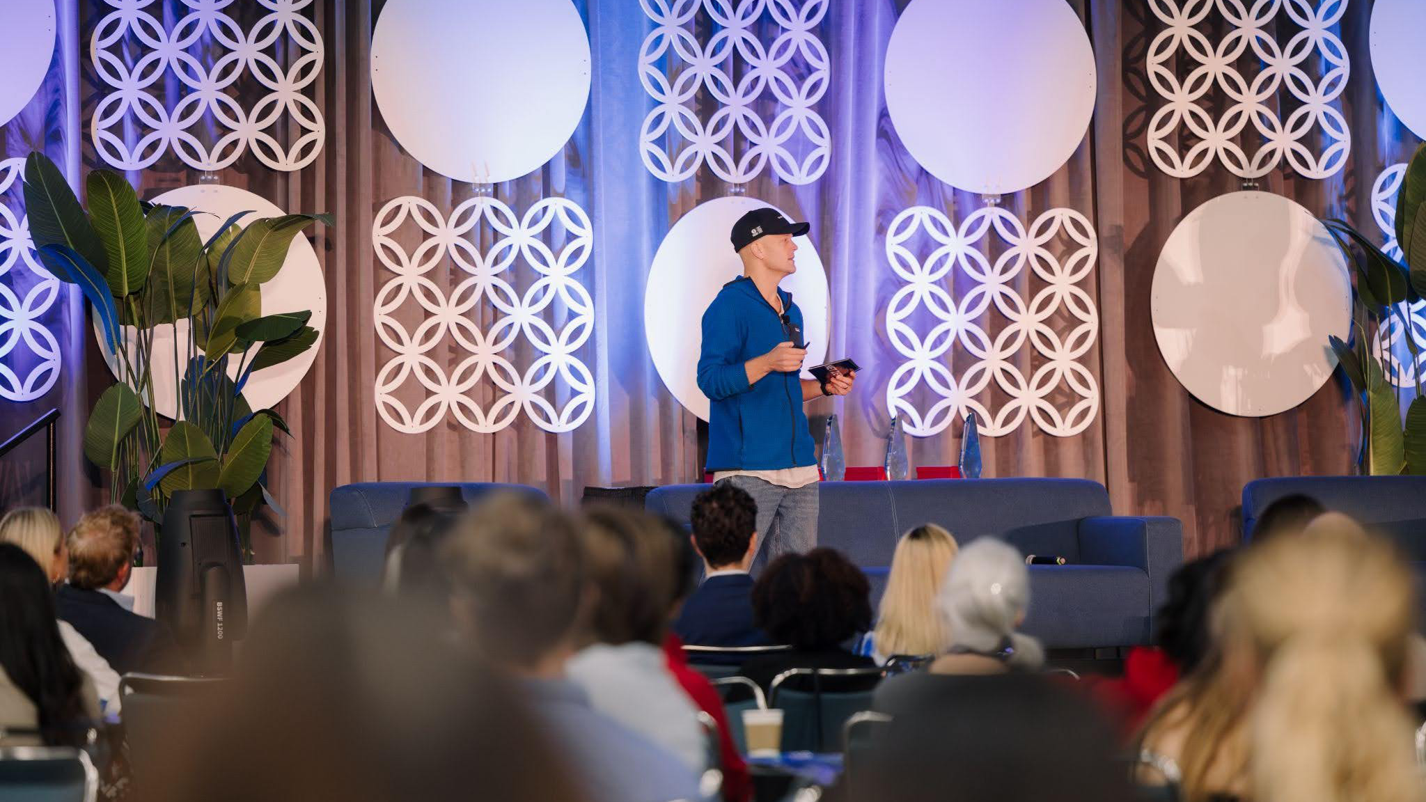 A man in a blue hoodie and baseball cap speaks on stage before an audience, with geometric wall decorations and plants in the background.