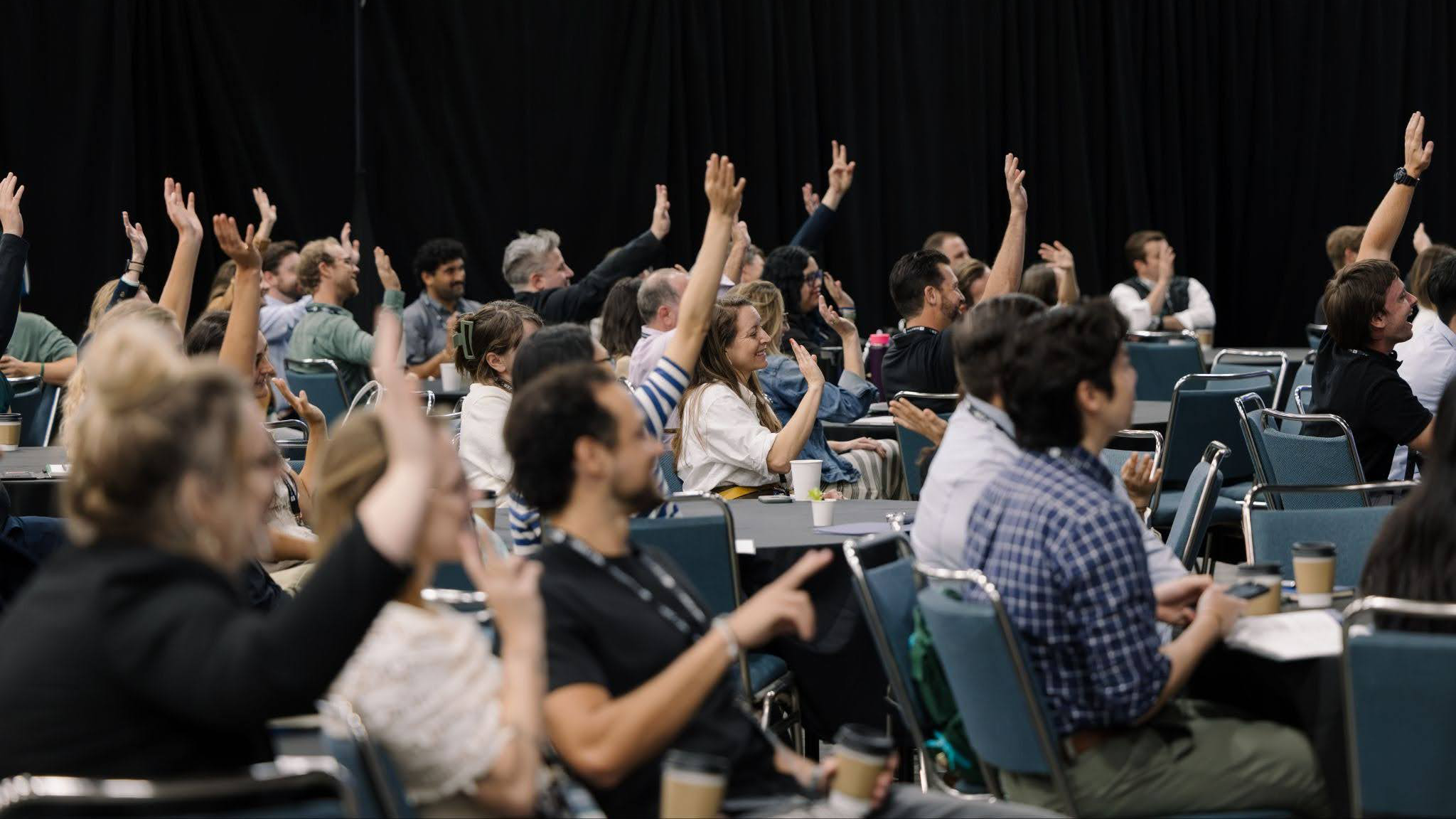 A group of people sitting at tables in a conference room with their hands raised.