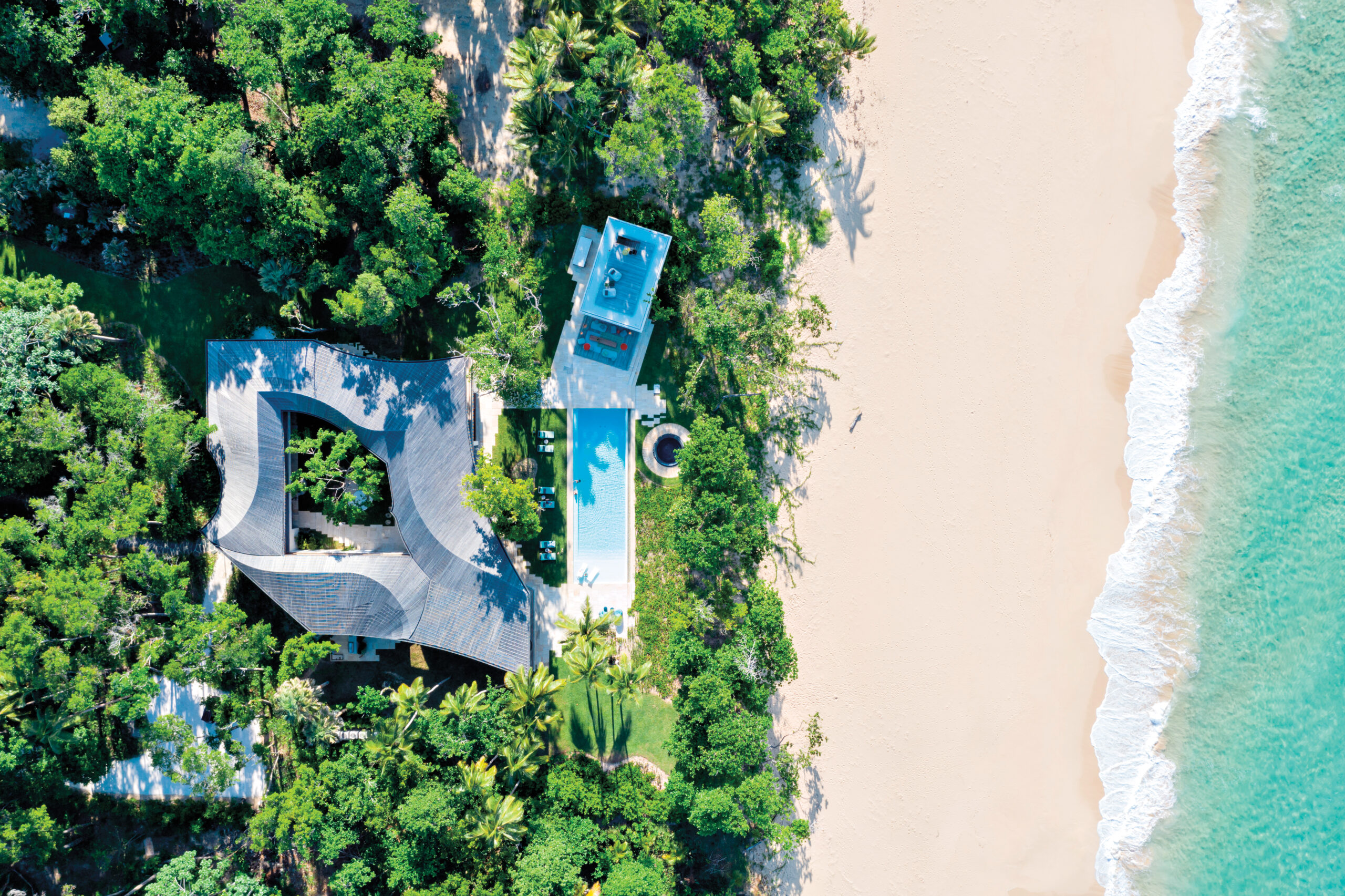 Birds-eye view of a beach with turquoise water, white sand and greenery, with a sculptural house in the forest.