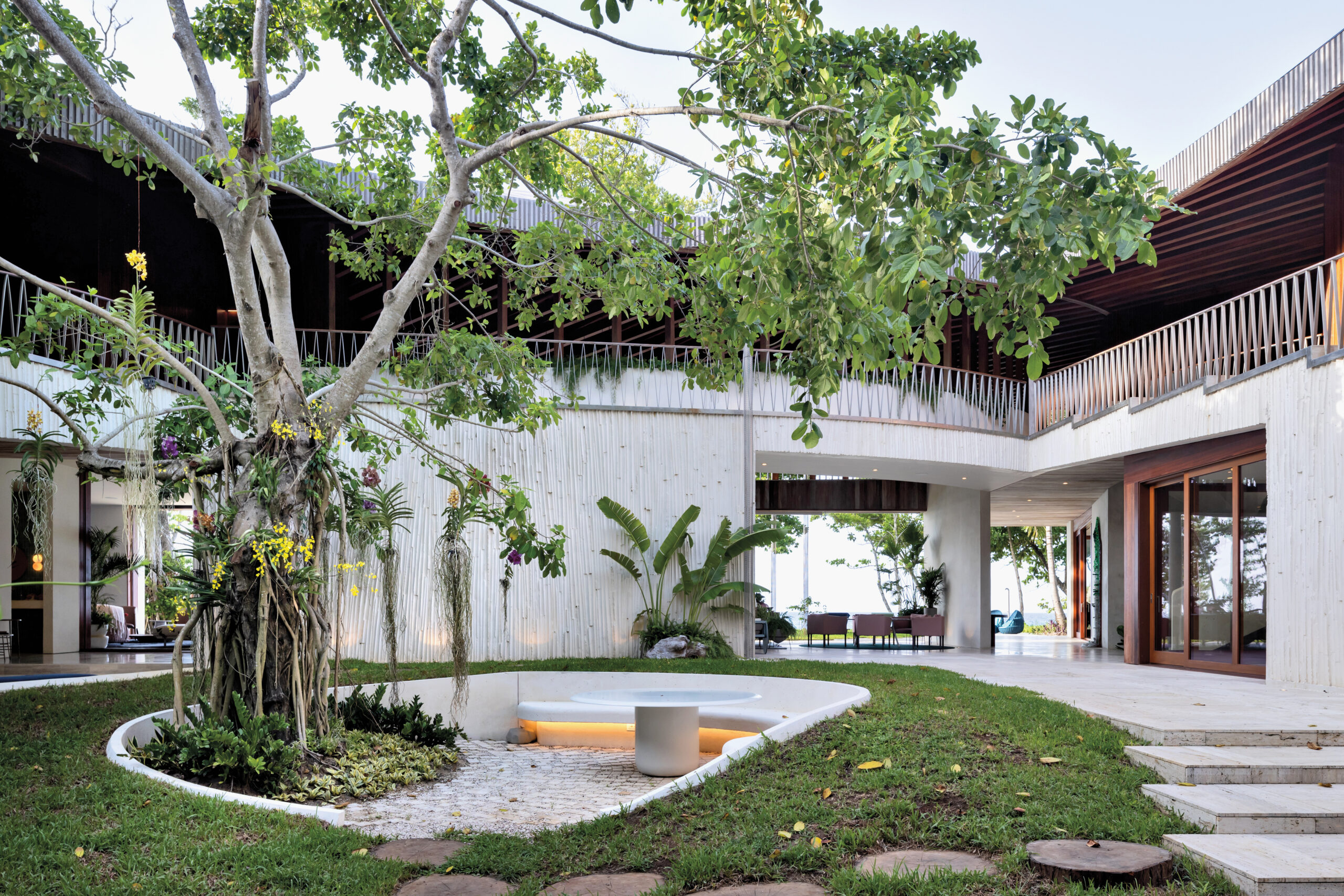 A modern residential building with an inner courtyard that features a sunken seating area and large tropical trees and plantings.